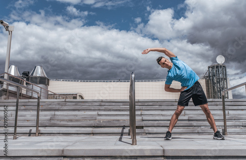 Fototapeta Naklejka Na Ścianę i Meble -  A male athlete in the summer in the city does stretching, preparing for jogging, training in the morning in the fresh air. Free space for motivation text. Youth lifestyle, active modern guy athlete.