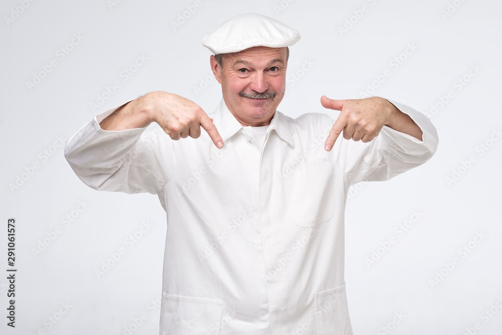 Smiling senior cook in white uniform smiling and pointing down trying to pay attention to important information.
