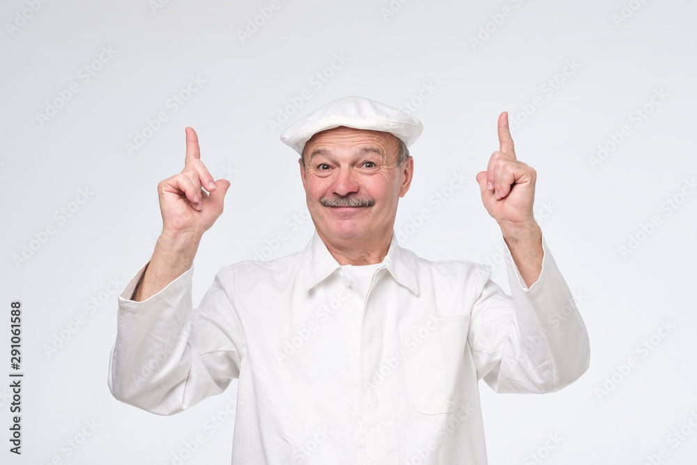 Smiling senior cook in white uniform smiling and pointing up trying to pay attention to important information. Smiling senior cook in white uniform smiling and pointing up trying to pay attention to important information.