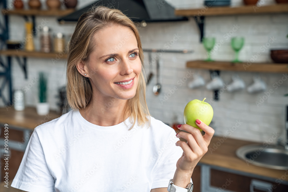 Close up of a beautiful blond who smiles and holds an apple in her hand. girl stands in the kitchen in a white t shirt Close up of a beautiful blond who smiles and holds an apple in her hand. girl stands in the kitchen in a white t shirt