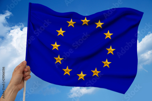 male hand holds against the background of the sky with clouds the national flag of the European Union on a luxury satin texture, silk with waves, closeup, copy space