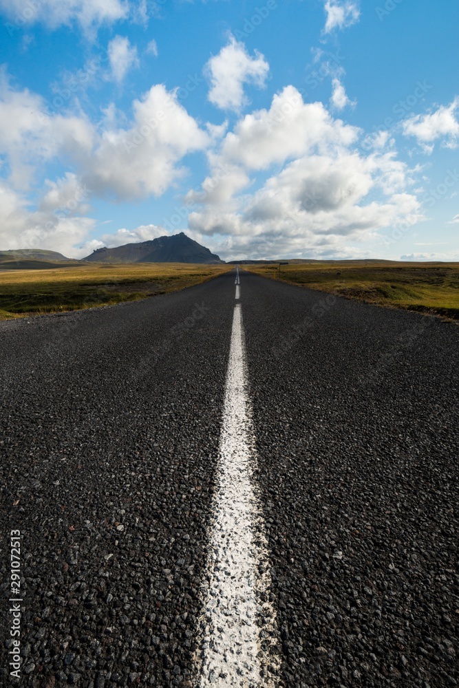 Low angle shot of an icelandic road extending to then horizon, with distant mountains and under a blue sky with puffy clouds