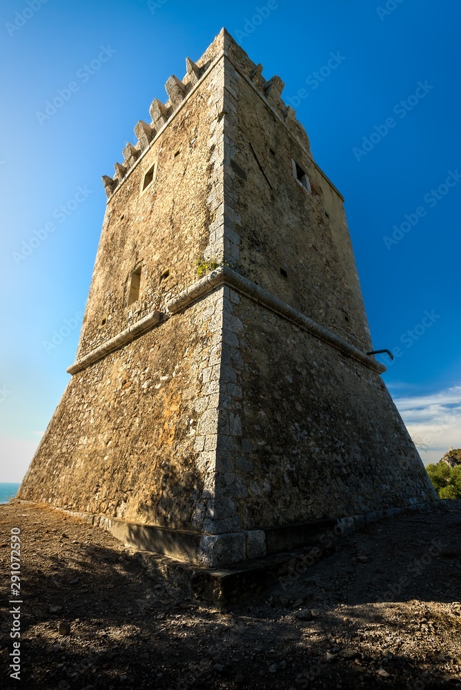 Symmetrical shot of an italian medieval stone watchtower with battlements on top, against a blue sky with distant clouds Symmetrical shot of an italian medieval stone watchtower with battlements on top, against a blue sky with distant clouds