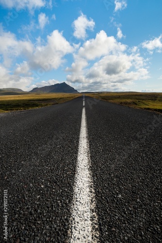 Low angle shot of an icelandic road extending to then horizon, with distant mountains and under a blue sky with puffy clouds