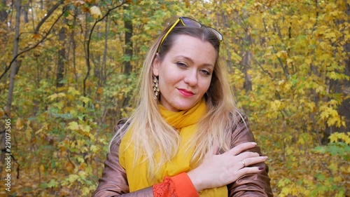 Pretty woman smiles in forest or park on background of trees with yellow leaves in autumn. Female in brown leather jacket with orange scarf and yellow glasses on her head