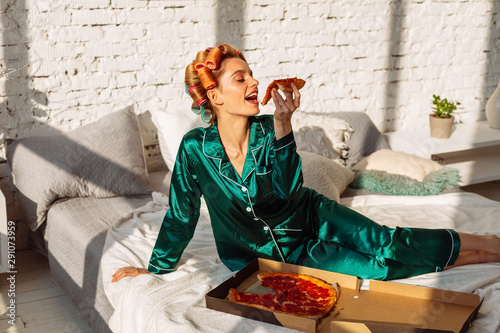 young cheerful woman in silk nightwear eating tasty pizza in bed, unhealthy eating