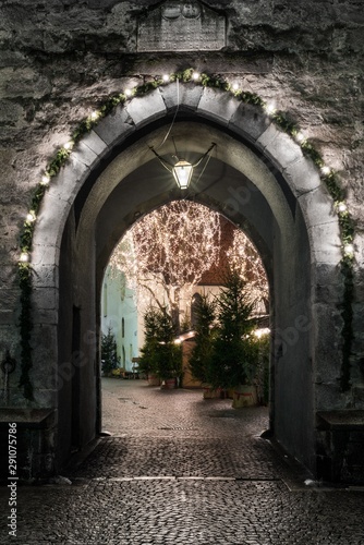 An ancient stone gate with a marble pointed arch decorated with christmas lights; beyond the gate there are two trees adorned with lights