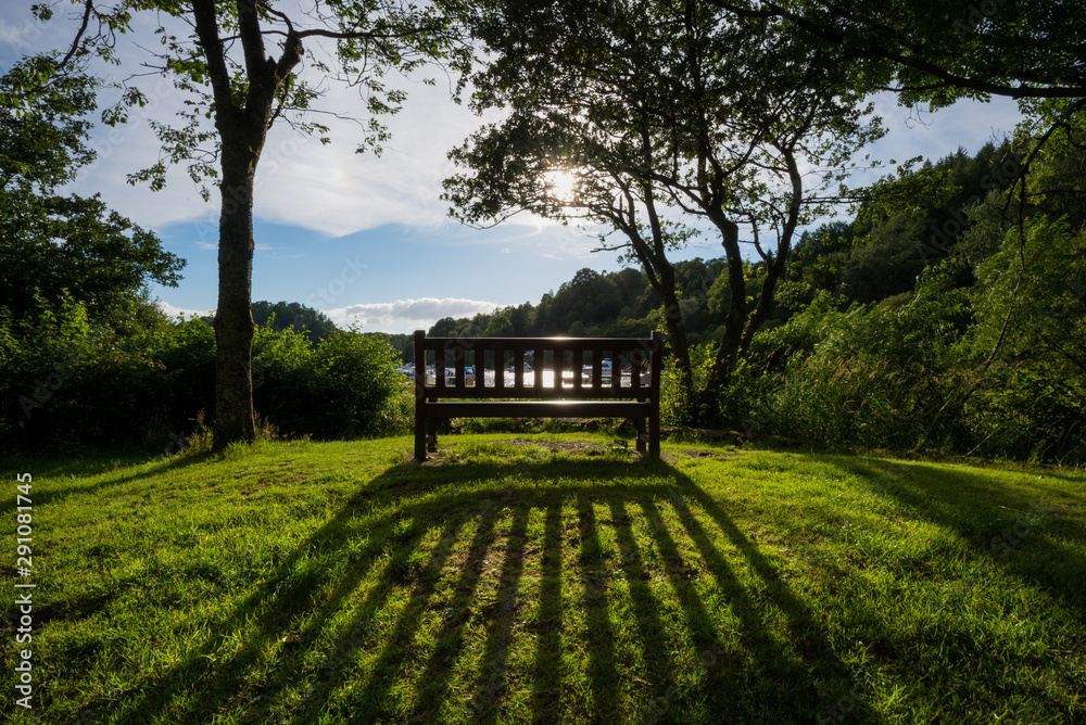 In a meadow overlooking a scottish lake at sunset, a wooden bench is casting an elongated shadow In a meadow overlooking a scottish lake at sunset, a wooden bench is casting an elongated shadow