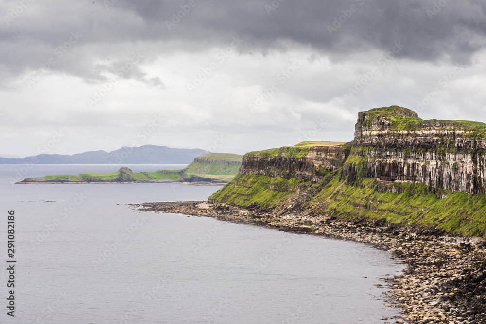 Scottish landscape with a shore covered with pebbles, the Kilt Rock cliff and distant mountains, under a cloudy sky