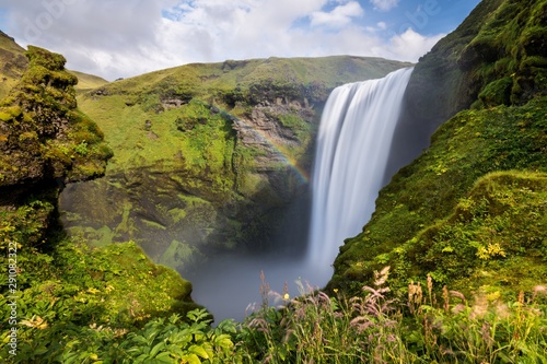 Long exposure of the icelandic waterfall of Skogafoss  surrounded by cliffs covered with green musk  under a blue sky with clouds