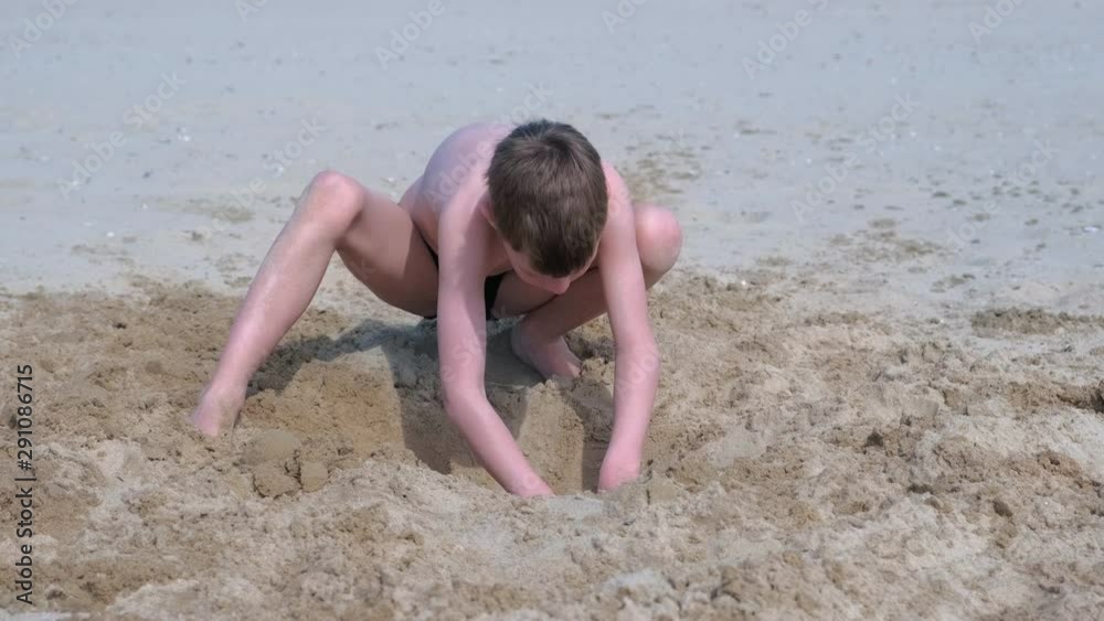 Child boy playing on sand beach digging shovel deep hole on summer vacation on sea ocean coast. Outdoor summer activities at vacation. Boy is sunbathing in swimming trunks.
