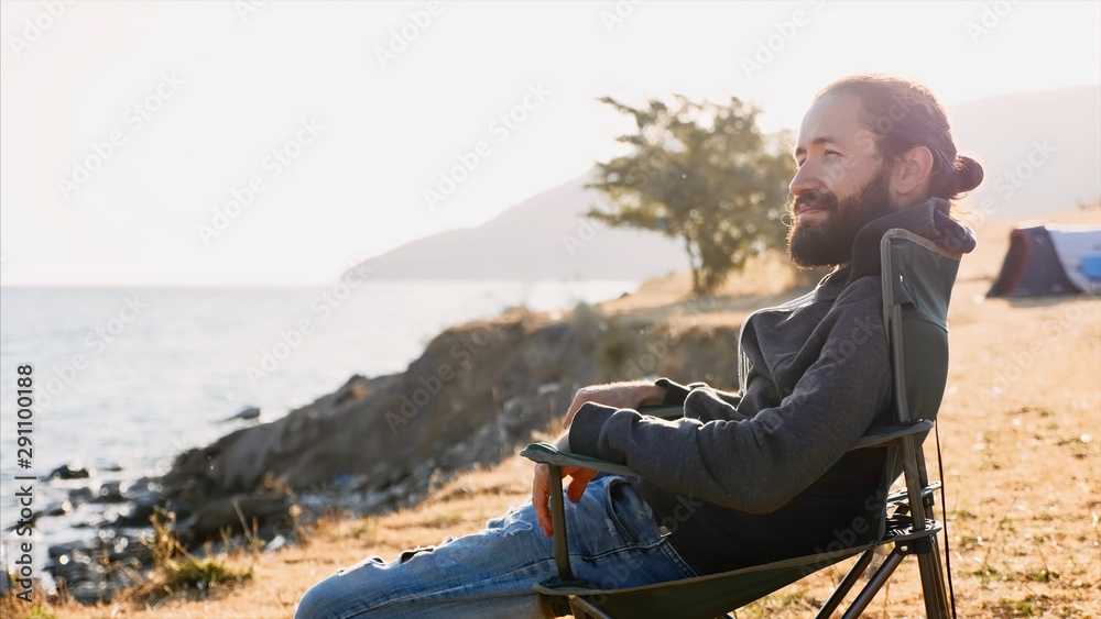 Young man sits on chair at campsite on lake shore