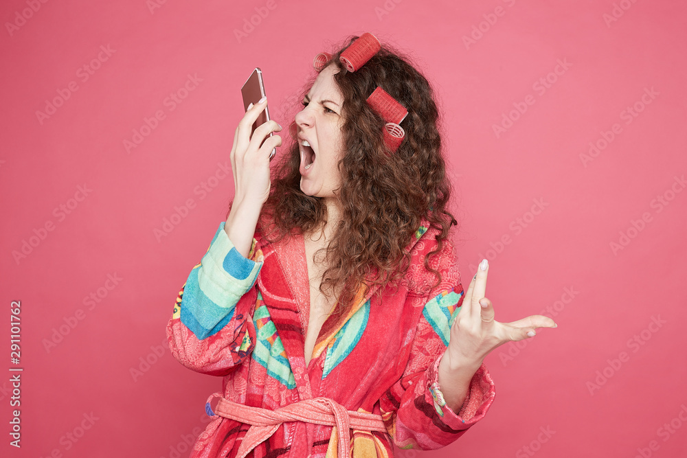 Desperate furious housewife gestures with annoyed face expression, talks on smart phone with husband, sorts out relationships, screams loudly, poses against pink studio wall. Negative human emotion. Desperate furious housewife gestures with annoyed face expression, talks on smart phone with husband, sorts out relationships, screams loudly, poses against pink studio wall. Negative human emotion.