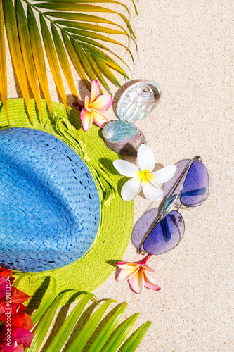 Blue and green straw hat with sunglasses, sea shells and frangipani flowers with green palm leafs on sand, summer vacation concept, top view, vertical composition