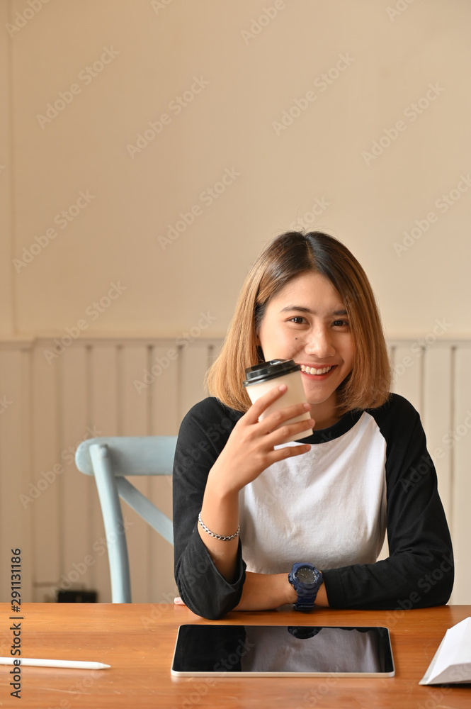 Young attractive woman holding coffee cup and looking at camera on creative table. Young attractive woman holding coffee cup and looking at camera on creative table.