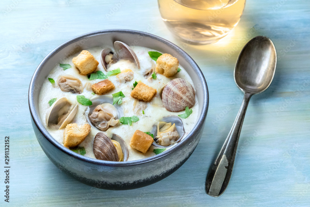 Clam chowder, decorated with fresh parsley and croutons, on a blue background, with a glass of white wine and a spoon Clam chowder, decorated with fresh parsley and croutons, on a blue background, with a glass of white wine and a spoon