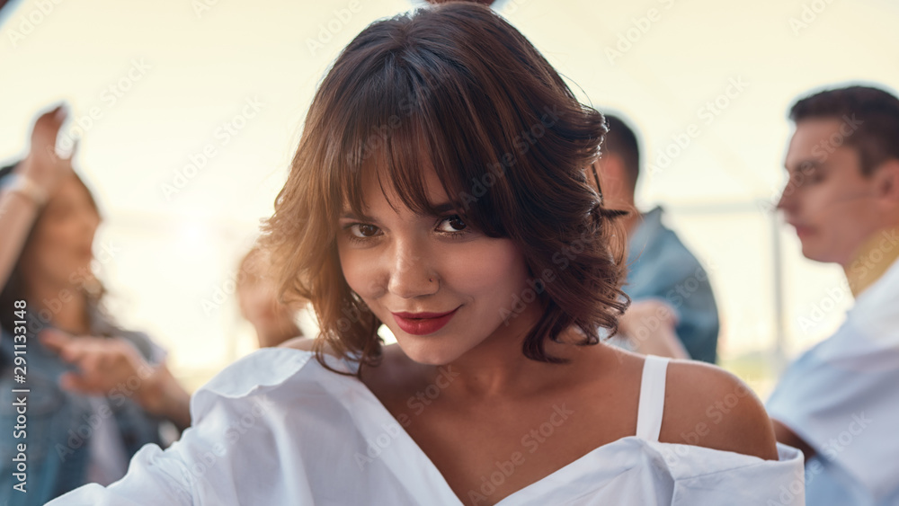 Flirty beauty. Pretty and young woman in white shirt is smiling and looking at camera while standing on rooftop terrace with her friends Flirty beauty. Pretty and young woman in white shirt is smiling and looking at camera while standing on rooftop terrace with her friends