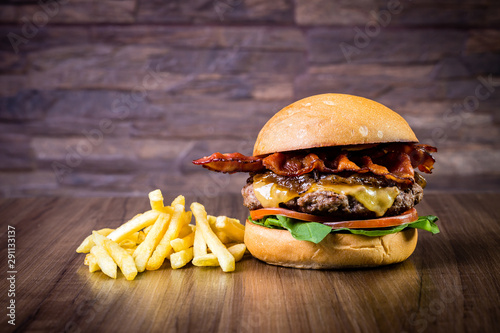 Craft beef burger with cheese, bacon, rocket leafs, caramelize onion and french fries on wood table and rustic background.