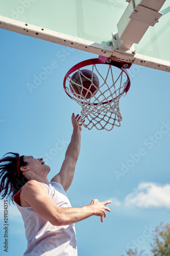 Image of athlete man throwing ball into basketball hoop on sports field on street on summer day.