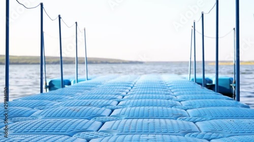 Blue floating plastic pontoon pier with rope railings rocking on waves at lake or river beach. Dock for small boats and people walkway