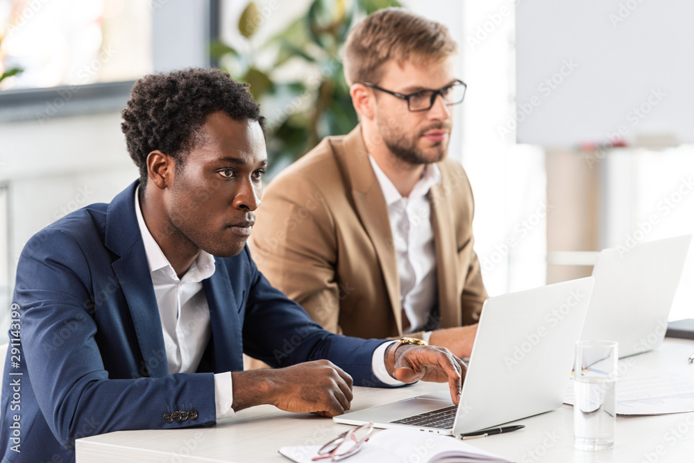 Obraz premium two multiethnic businessmen using laptops at table in office