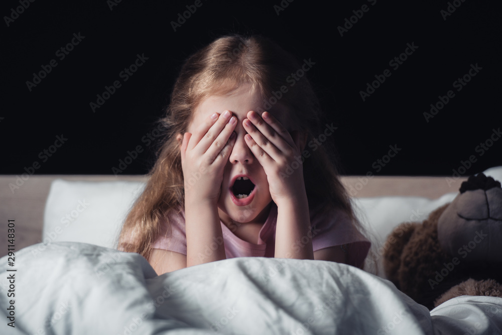 frightened kid screaming and covering eyes with hands while sitting on bedding near teddy bear isolated on black frightened kid screaming and covering eyes with hands while sitting on bedding near teddy bear isolated on black