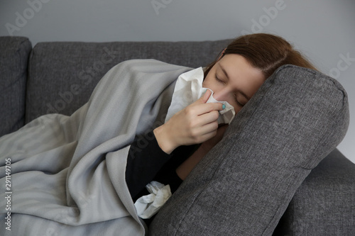 Young woman on the sofa blowing nose into a white paper tissue. Studio shot of young woman with allergy symptoms sneezing into a tissue. Flu, cold or allergy symptom.  photo