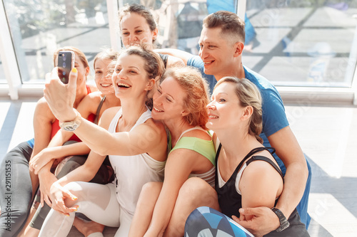 Group of positive young people take a selfie on a smartphone after a group of yoga classes in their bright room with a large window. Concept lovers of yoga and pilates