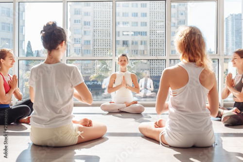 Stylish young women meditate in padmasana sitting on the floor in a bright gym with large windows. Concept of maintaining of freshness of mind and eliminating lethargy and drowsiness, sports unity photo