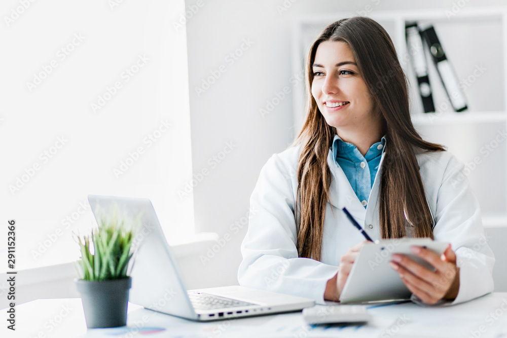 Smiling young female doctor working at the clinic , she is using a computer and writing medical reports. Smiling young female doctor working at the clinic , she is using a computer and writing medical reports.