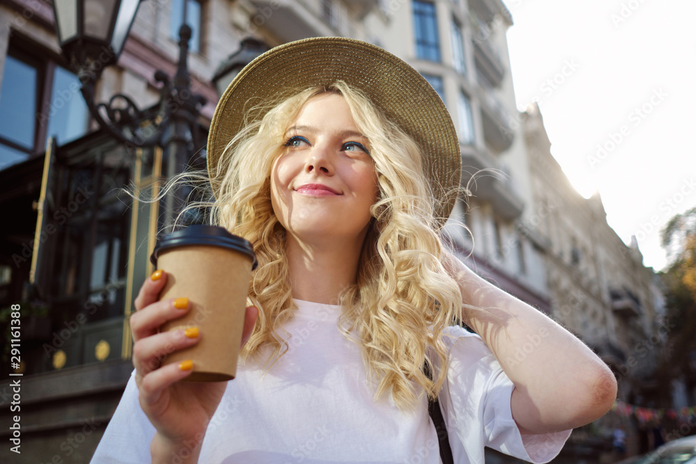 Portrait of pretty smiling blond girl in hat dreamily looking away with coffee to go on city street