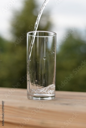 A stream of water flowing into an empty transparent glass standing on a wooden board.
