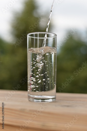 A jet of water flowing into a transparent glass with water and air bubbles standing on a wooden board.