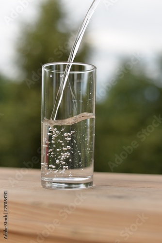 A jet of water flowing into a transparent glass with water and air bubbles standing on a wooden board.