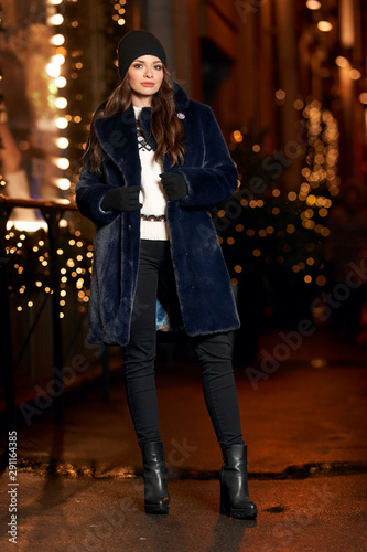 Pretty girl in blue fur coat standing and posing at christmas decorated city street at night. Beautiful lighs bokeh
