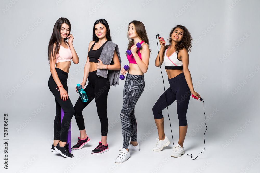 Fototapeta premium Group portrait of young sporty excited beautiful girls with exercise equipment standing beside white wall laughing and talking together. Candid funny students waiting 