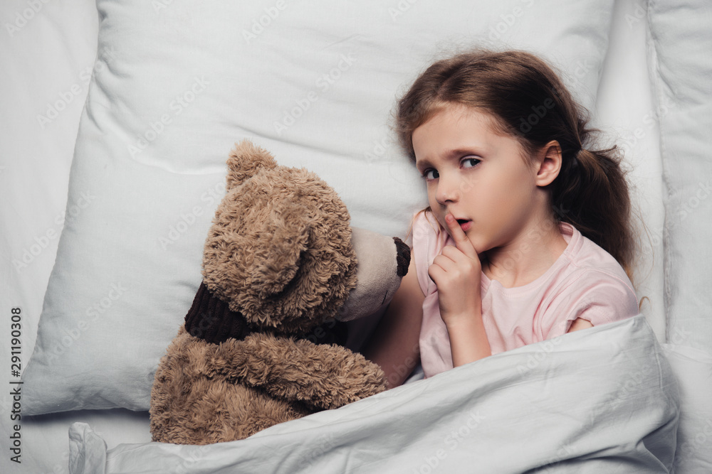 top view of scared child showing hush sign and looking at camera while lying in bed with teddy bear top view of scared child showing hush sign and looking at camera while lying in bed with teddy bear