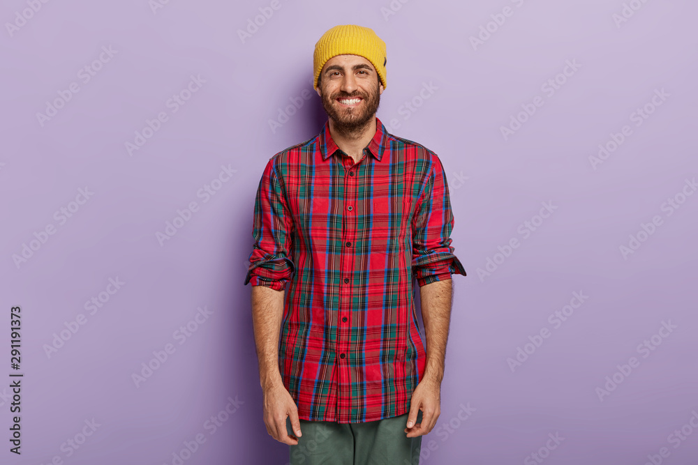 Portrait of happy guy with bright smile, stubble, wears yellow hat and checkered shirt, enjoys free time for walk with girlfriend, isolated over purple wall looks straightly at camera. Youth and style