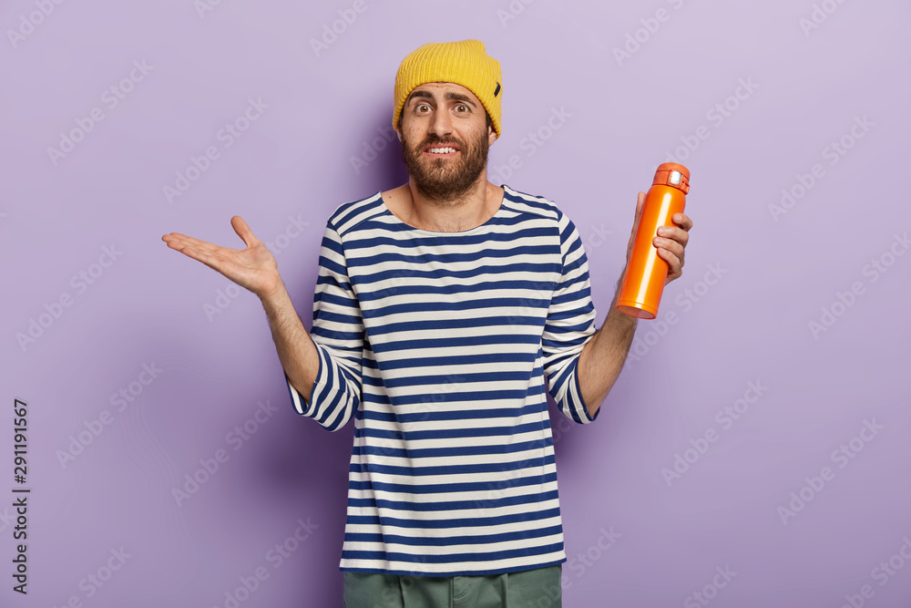 Unaware confused Caucasian man raises palm with hesitation, holds orange flask with hot drink, wears yellow hat and striped jumper, doesnt know what to do isolated over purple wall. Tourist with drink Unaware confused Caucasian man raises palm with hesitation, holds orange flask with hot drink, wears yellow hat and striped jumper, doesnt know what to do isolated over purple wall. Tourist with drink