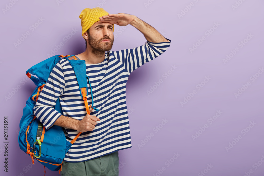 Serious handsome man tries to see something into distance, keeps palms near forehead, carries big tourist rucksack with personal things, stands against purple background, blank space for promo