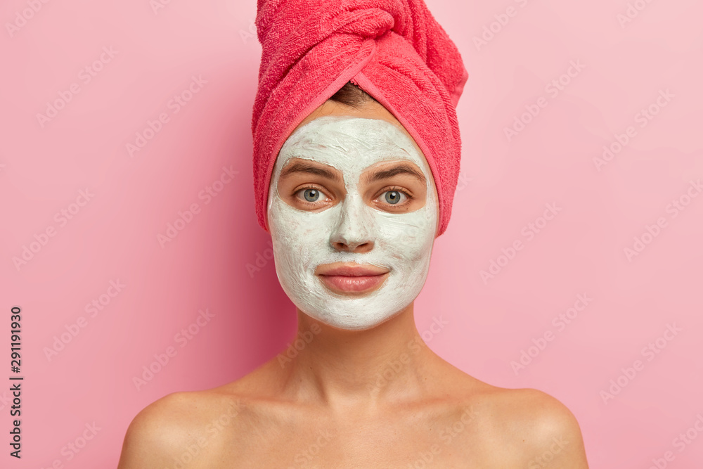 Close up shot of serious young woman with clay facial mask, wears wrapped towel, nourishes skin with vitamins, looks straightly at camera, stands naked against pink background, pampers complexion Close up shot of serious young woman with clay facial mask, wears wrapped towel, nourishes skin with vitamins, looks straightly at camera, stands naked against pink background, pampers complexion