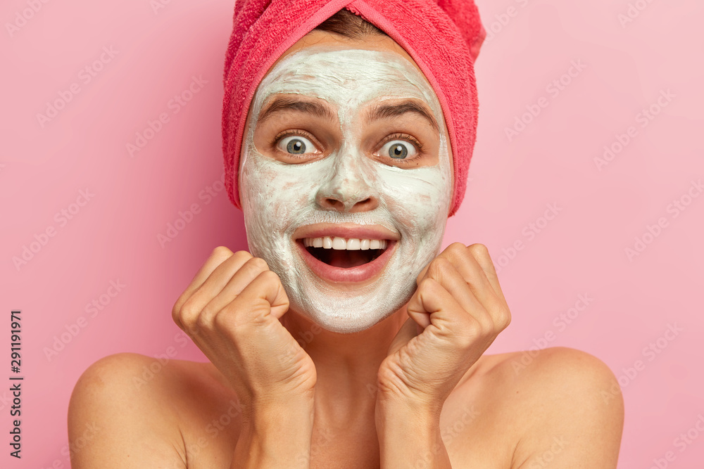 Φωτογραφία Close up portrait of happy young Caucasian woman smiles broadly, keeps both hands under chin, cannot believe her eyes, applies clay mask, has joyful face expression, poses over pink background