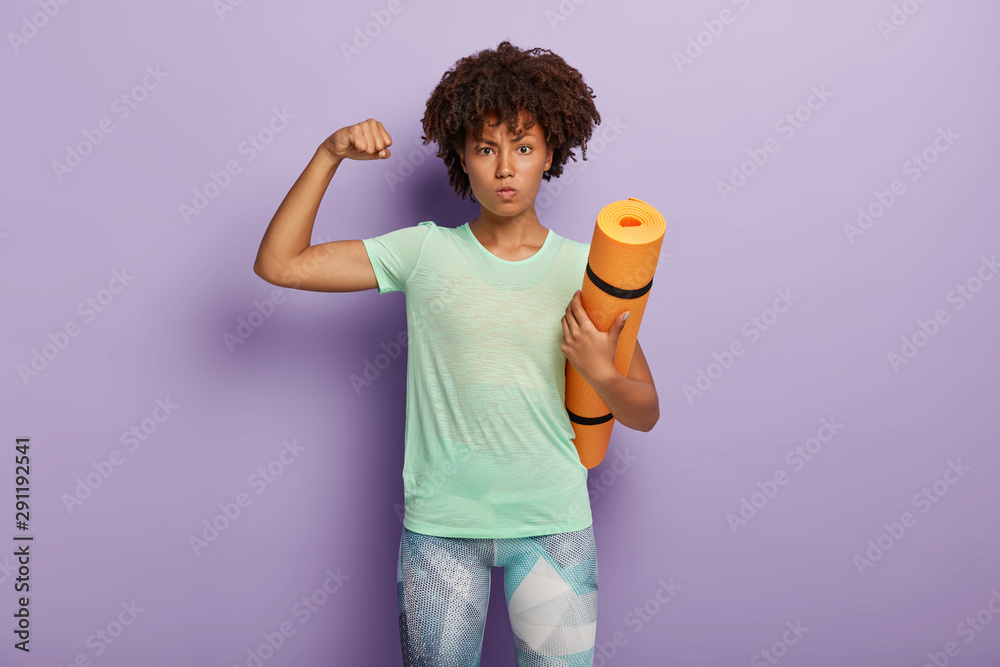 Powerful strong African American woman raises arm, shows biceps, holds fitness mat for gym training, dressed in active wear, isolated over purple background, ready for workout or yoga practice Powerful strong African American woman raises arm, shows biceps, holds fitness mat for gym training, dressed in active wear, isolated over purple background, ready for workout or yoga practice