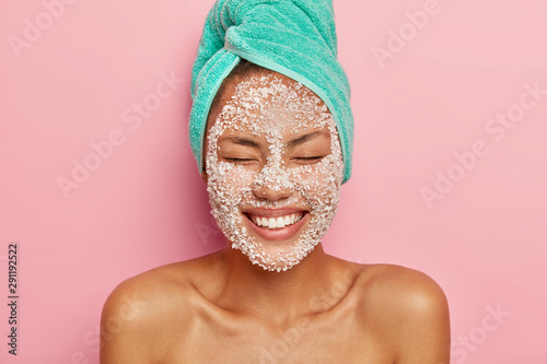 Headshot of pretty smiling woman applies salt granules on face, keeps eyes closed, shows white perfect teeth, wears turquoise towel, poses shirtless against pink background, enjoys effective result