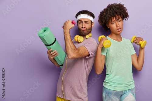 Tired African American woman raises two dumbbells, works on biceps, determined unshaven man carries karemat for gymnastic training, stand backs, isolated over purple background. Bodybuilding