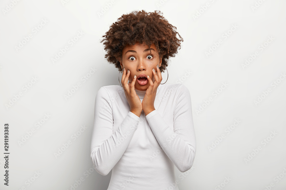 Photo of frightened curly haired woman gasps from fear, keeps hands on cheeks, cannot believe in shocking news, wears poloneck sweater, isolated on white background. Human facial expressions Photo of frightened curly haired woman gasps from fear, keeps hands on cheeks, cannot believe in shocking news, wears poloneck sweater, isolated on white background. Human facial expressions