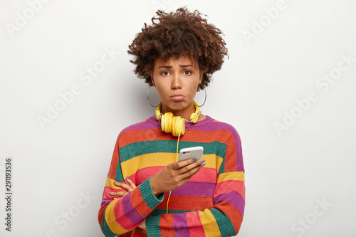 Dissatisfied teenager with curly hairstyle, uses smart phone, feels lonely and upset, dressed in striped sweater, looks at camera with regret and sadness, isolated, upset boyfriend doesnt answer