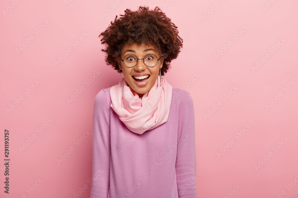 Isolated shot of pretty curly woman expresses joy, enjoys funny moment, laughs at something, smiles positively, wears round spectacles, purple casual wear, isolated on pink background. Happiness Isolated shot of pretty curly woman expresses joy, enjoys funny moment, laughs at something, smiles positively, wears round spectacles, purple casual wear, isolated on pink background. Happiness