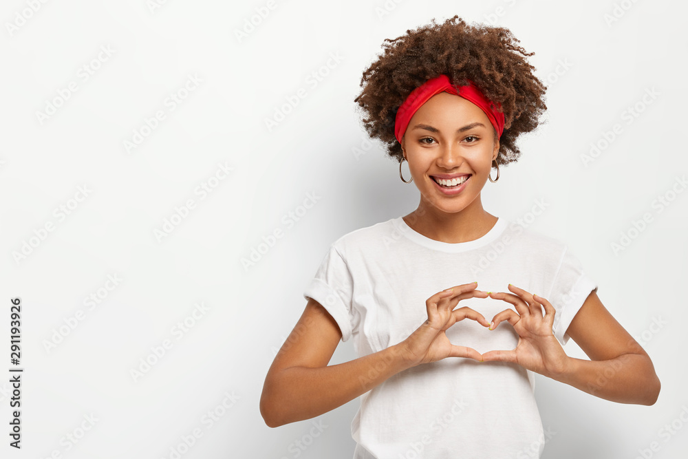 Love to you, my dear. Glad satisfied African American woman makes heart gesture, smiles pleasantly at camera, wears casual outfit, expresses good feelings to lover, poses indoor over white background - obrazy, fototapety, plakaty 