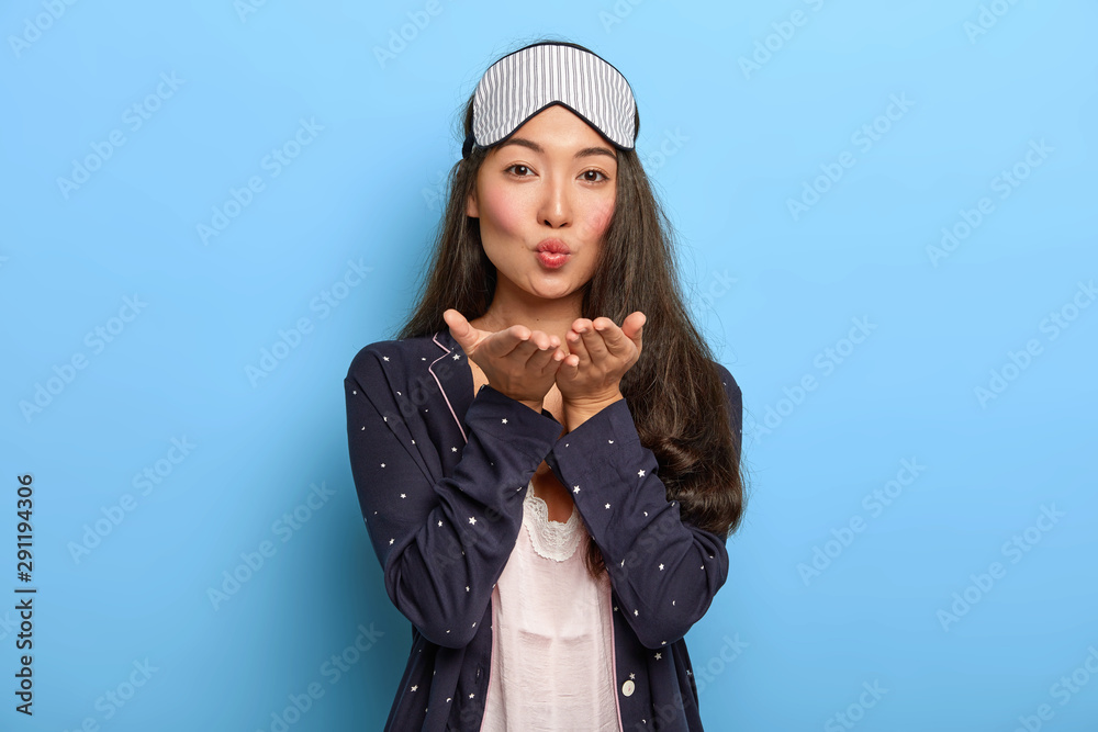 Portrait of lovable brunette Asain woman sends air kiss to husband, wakes up in morning, wears sleep mask and night costume, rests at home, poses against blue wall. Lady dressed in homewear. Portrait of lovable brunette Asain woman sends air kiss to husband, wakes up in morning, wears sleep mask and night costume, rests at home, poses against blue wall. Lady dressed in homewear.